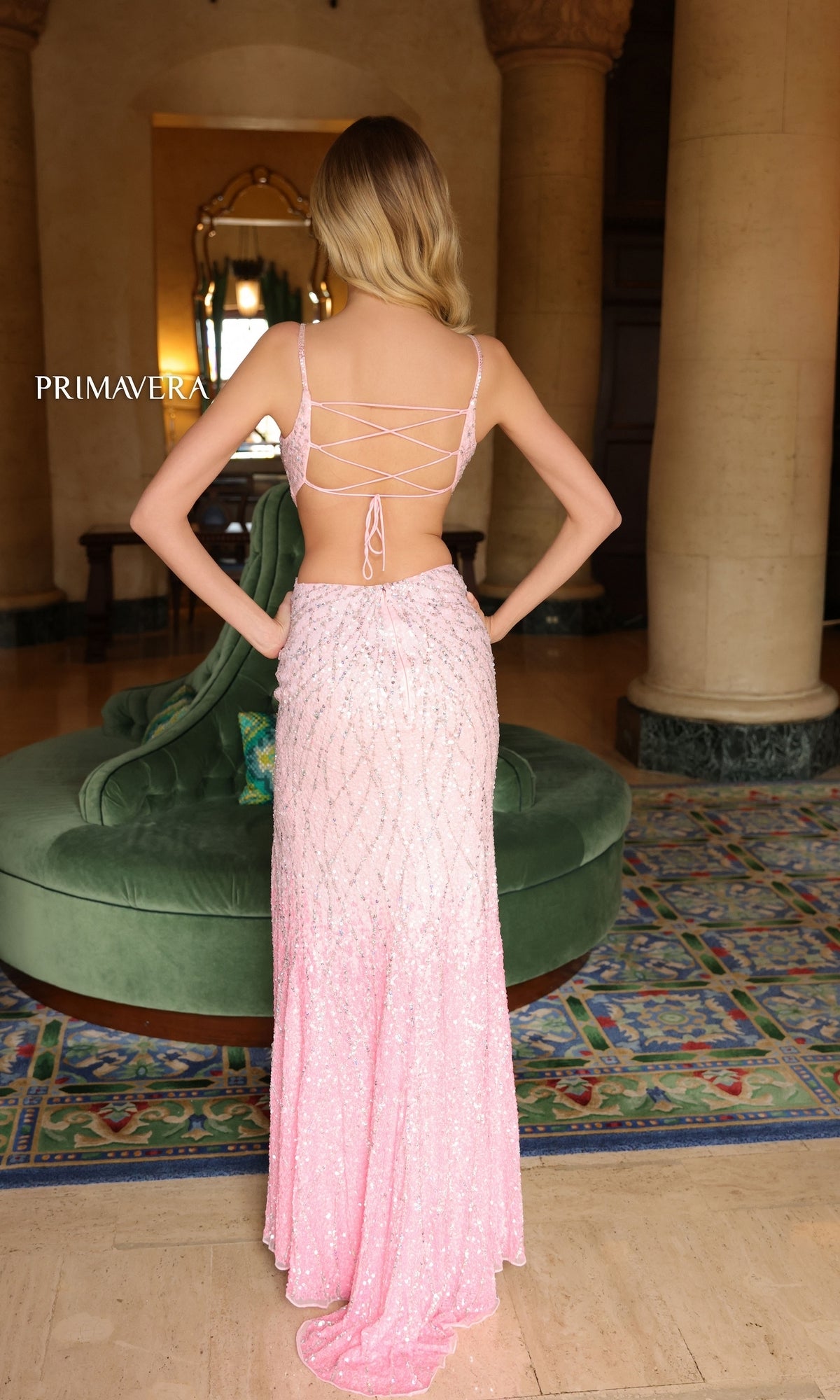 A woman stands indoors facing away, wearing the Primavera Ombre-Beaded Cut-Out Prom Dress 4136—a floor-length, light pink gown with beads, an open back, and lace-up detail.
